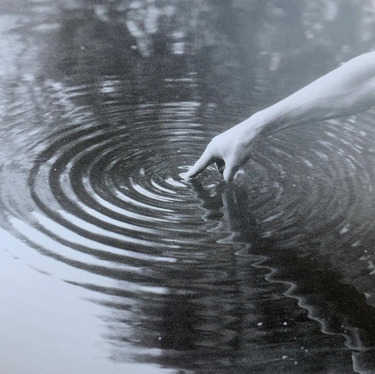 Black-and-white photograph of a hand reaching down to touch dark water, with rings rippling outward.
