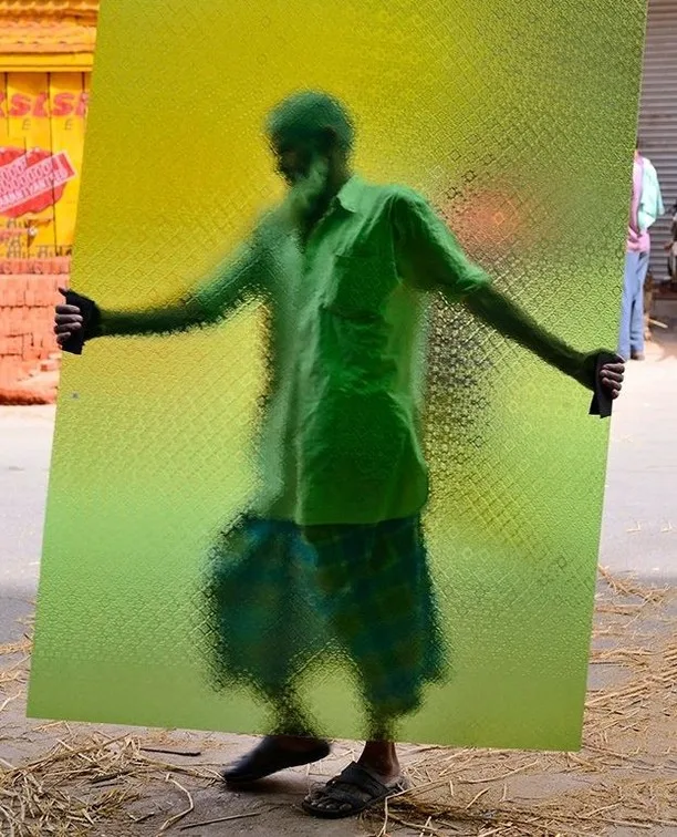 A man carries a large pane of green patterned glass in the street; he is blurred behind it and the glass tints him and the background green and yellow.