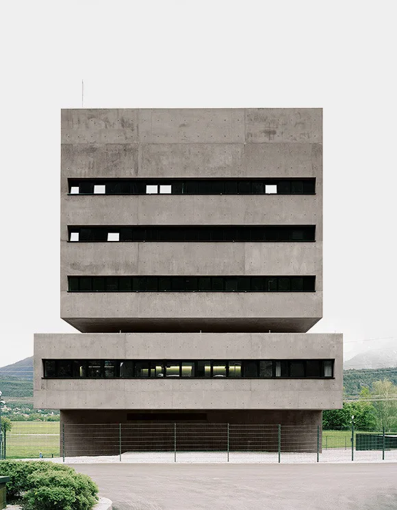 A grey concrete building of two stacked blocks with long, narrow, dark windows, seen from the front under a pale sky, with a green field behind.