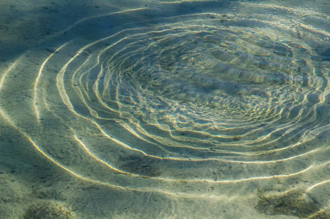 Concentric rings of light and ripples on a sandy seabed, seen through clear, shallow water.